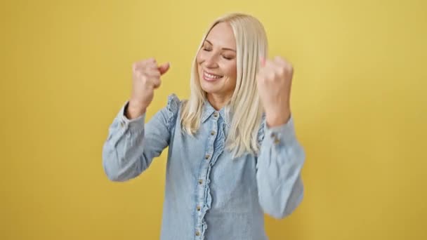 Triumfalna młoda blondynka odstawia zwycięską pozę w jeansowej koszuli - stojąc, podnosząc ramiona, ciesząc się z sukcesu, świętując na żółtym, odosobnionym tle! Co za zwycięstwo.! - Materiał filmowy, wideo