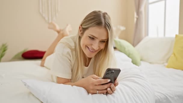 Здивована, красива молода блондинка лежить в ліжку, використовуючи свій смартфон у затишній кімнаті. смс її друзів вранці, приваблива жінка в піжамах прокидається, щоб здивувати текст. - Кадри, відео