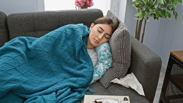 Femme hispanique dormant sur un canapé sous une couverture bleue dans un salon confortable - Photo, image