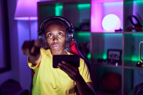 Femme afro-américaine jouant à des jeux vidéo avec smartphone pointant du doigt la caméra et à vous, geste confiant regardant sérieux  - Photo, image