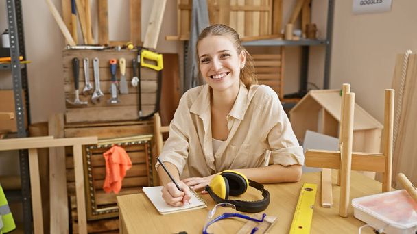 Eine lächelnde junge Frau skizziert in einer Tischlerei, umgeben von Werkzeug und Holz. - Foto, Bild
