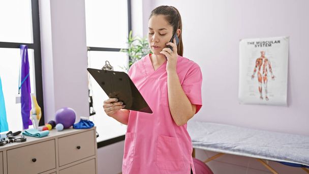 Una mujer enfocada en una bata rosa habla por teléfono mientras sostiene un portapapeles en la sala de tratamiento de una clínica de fisioterapia. - Foto, Imagen