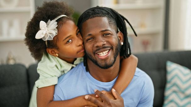 Αφροαμερικάνος πατέρας και κόρη χαμογελούν με αυτοπεποίθηση αγκαλιάζοντας ο ένας τον άλλον καθισμένοι στον καναπέ στο σπίτι - Φωτογραφία, εικόνα