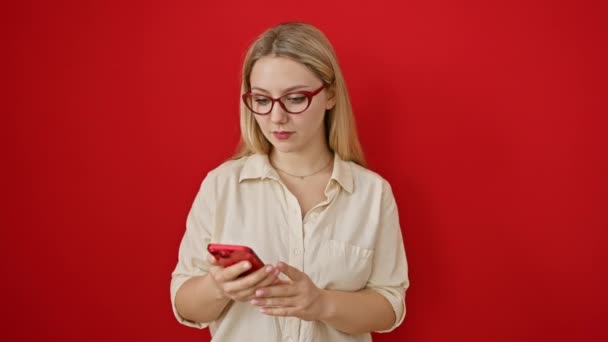 Vrolijke, zelfverzekerde jonge blonde vrouw geeft duimen-up, sportieve een succesvolle glimlach tijdens het sms 'en op de smartphone, het uiten van puur geluk over geïsoleerde rode muur pittige moderne meisje' s levensstijl - Video