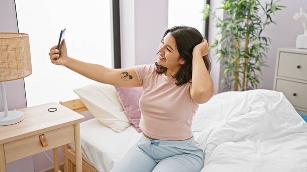 Sorrindo jovem hispânica tomando uma selfie em seu quarto bem iluminado e acolhedor, adicionando um toque de estilo de vida e positividade. - Foto, Imagem