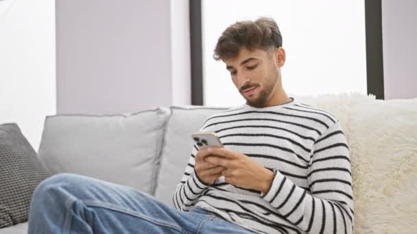 Jovem árabe alegre homem alegremente mensagens de texto em seu smartphone enquanto sentado confortavelmente em seu sofá, exalando a felicidade na calorosa e aconchegante sala de estar de sua casa - Filmagem, Vídeo