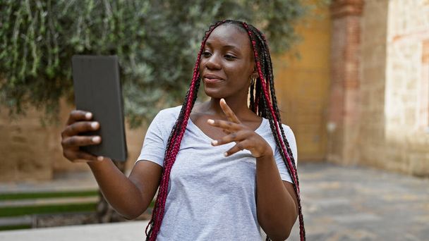 Mulher americana africana bonita repleta de confiança, sorrindo alegremente enquanto tem uma chamada de vídeo agradável e divertida em seu gadget em uma rua da cidade sob a luz solar urbana quente - Foto, Imagem