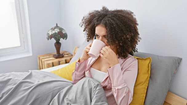 Mulher hispânica com cabelo encaracolado, vestindo pijama, tomando café em um ambiente acolhedor quarto, olhando relaxado. - Foto, Imagem