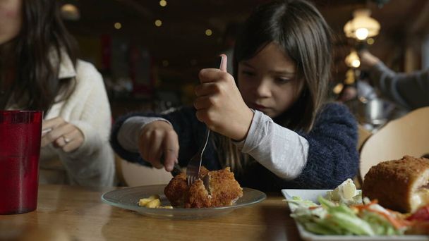 Niña comiendo comida en el restaurante, niño disfrutando de cordon bleu pollo para la hora de comer en el acogedor interior de madera. Familia en el restaurante - Foto, imagen
