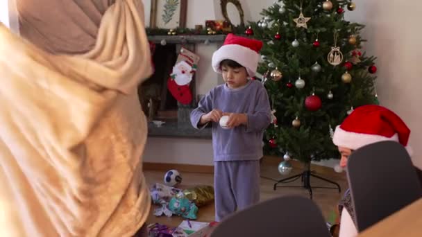 Rodzina gromadzi się wokół chłopca w czerwonym kapeluszu w Boże Narodzenie rano, rozpakowywanie prezentów - Materiał filmowy, wideo