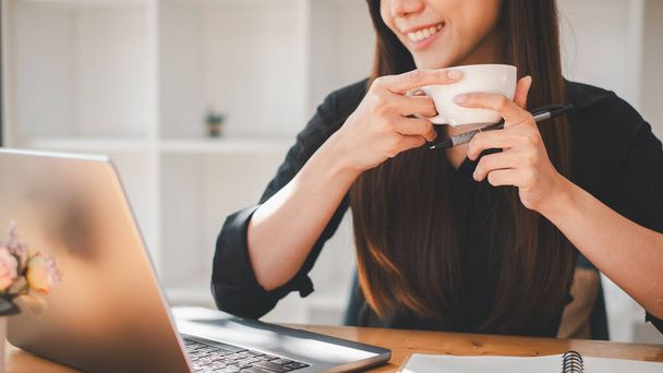 Neşeli genç bir kadın, parlak bir ofis ortamında laptopuyla çalışırken rahatlatıcı bir kahve molası verir. - Fotoğraf, Görsel