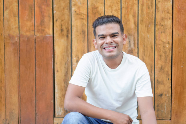 Junger Mann mit Zahnspange lächelt und sitzt lässig an einer Holzlattenwand. - Foto, Bild