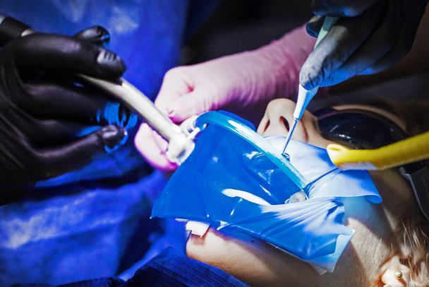 дантист и ассистент в перчатках лечат зубы подростка. Стоматологическая помощь и здоровье - Фото, изображение
