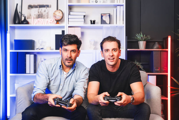 Νικητές και ηττημένοι παίκτες φίλων gamers που παίζουν βιντεοπαιχνίδια στην τηλεόραση χρησιμοποιώντας joysticks στο στούντιο με νέον μπλε φως. Comfy ζουν σε εσωτερικούς χώρους στο σπίτι με χαρούμενα νικητής αγωνίζονται. Πωλητέα. - Φωτογραφία, εικόνα