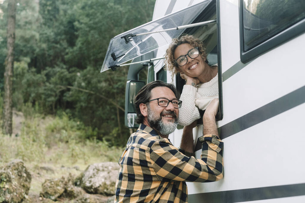 Boldog felnőtt pár élvezni úticél lakókocsi lakókocsi van Vanlife életmód. Nő az ablaknál bent, férfi kint zöld erdőkkel a háttérben. Alternatív élet nyaralás - Fotó, kép