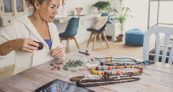  busy woman at home start new small business doing jewelry with beads and using laptop to sell on line or learn from tutorial. Indoor hobby leisure activity female people at the table alone - Foto, imagen