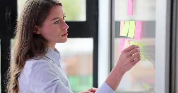 Businesswoman pasting sticker on a glass wall in office. Female employee looking at stickers indoors and planning and business to-do list - Footage, Video