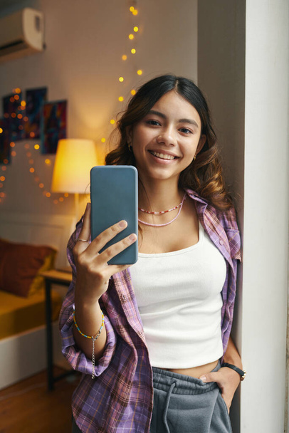 Portré vidám tizenéves lány loungewear áll a hálószoba ablakában, amikor selfie - Fotó, kép