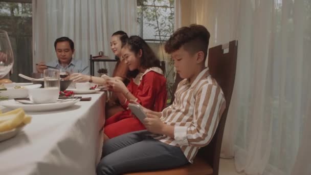 Boční pohled na sourozence, kteří hrají hry na chytrých telefonech, zatímco ostatní členové rodiny mluví během večeře - Záběry, video