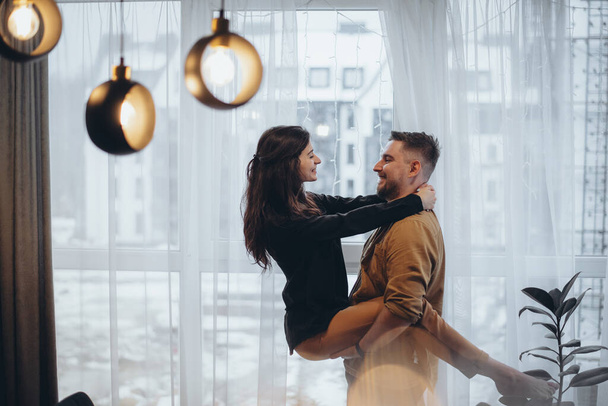 Young couple in a hotel near the window. The guy holds the girl in his arms. The girl holds her hands on the guy's neck. They smile. The city is blurry in the background. Glare in the foreground - Photo, Image