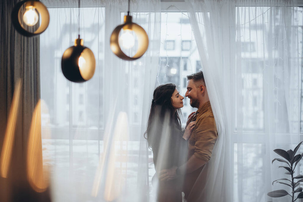 Ein junges Paar in einem Hotel steht am Fenster, umarmt und schaut einander an, in Vorhänge gehüllt. Im Hintergrund verschwimmt die Stadt. Im Vordergrund ein Kronleuchter mit Glühbirne und grellem Licht - Foto, Bild