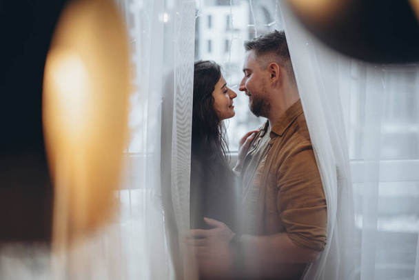 A young couple stands near the window, hugging and looking at each other, wrapped in curtains. The city is blurry in the background. In the foreground is a chandelier with a light bulb and glare - Photo, Image