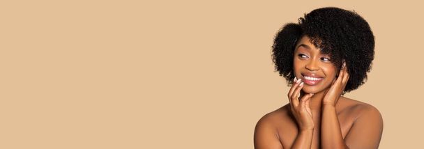 Radiant African American woman with natural afro hair smiling warmly, touching her face gently with both hands, looking aside at free space against beige background, panorama - Photo, Image