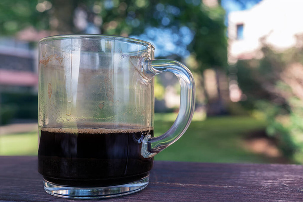 ein Glasbecher mit fertigem Kaffee, steht auf einem Holztisch im Park - Foto, Bild