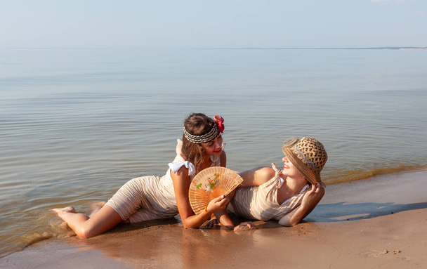 Dos mujeres jóvenes en trajes de baño retro, sombrero y abanico disfrutando de la naturaleza y unos a otros en la orilla de un mar tranquilo en un día de verano - Foto, imagen