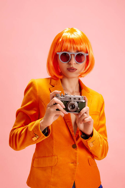 Żywa moda. Młoda kobieta w modnych okularach przeciwsłonecznych ubrana w pomarańczową marynarkę, z peruką patrzącą w kamerę i trzymającą aparat fotograficzny. Pojęcie osobistego wyrazu i pewności siebie poprzez styl. - Zdjęcie, obraz