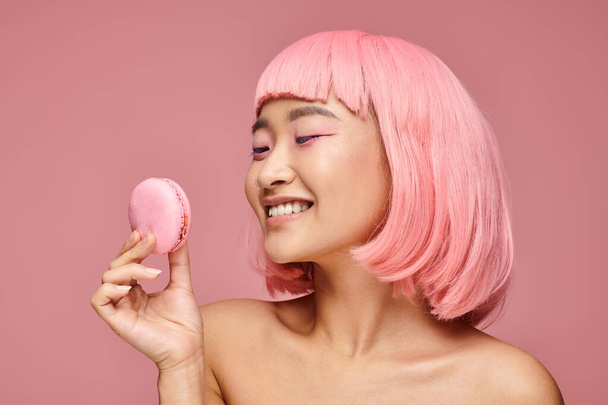 pretty asian woman in 20s looking to macaroon and smiling against vibrant background - Foto, Imagen