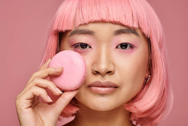 cerrar atractivo asiático mujer con rosa pelo posando con macaroon en vibrante fondo - Foto, Imagen