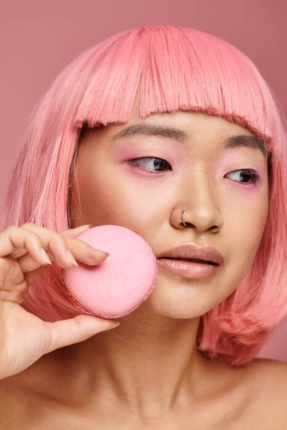 cerrar hasta asiático mujer con rosa pelo posando con macaroon y buscando a lado en vibrante fondo - Foto, imagen