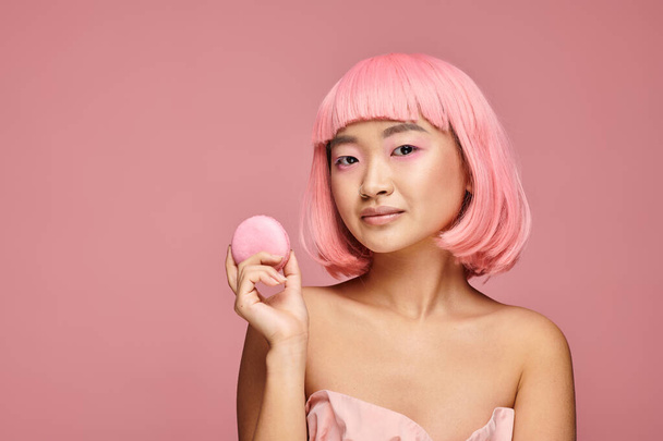 pretty asian young woman with pink hair and makeup holding macaroon with hand in vibrant background - Photo, Image