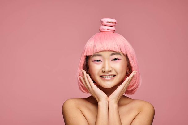 happy asian young woman with pink hair, hands and macaroons on head on vibrant background - Fotografie, Obrázek