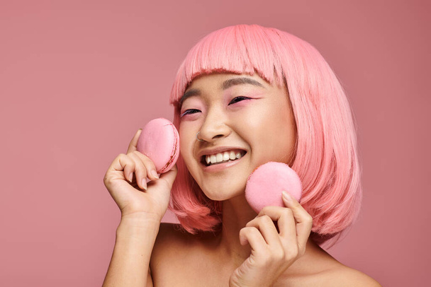 happy asian girl with pink hair laughing and holding macaroons with hand in vibrant background - Photo, Image