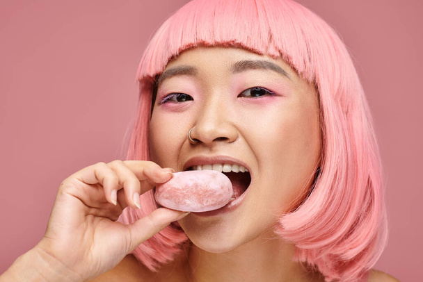 portrait of happy asian young woman with pink hair eating mochi against vibrant background - Photo, Image