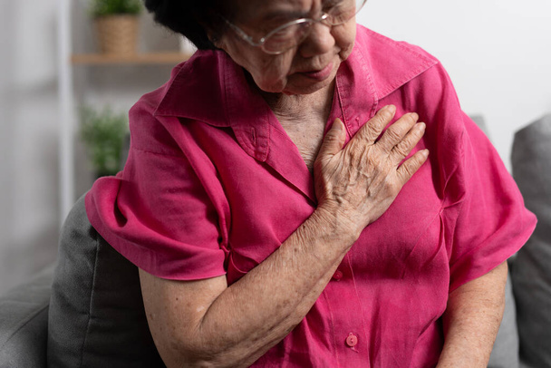 Ασιάτισσα ηλικιωμένη γιαγιά που υποφέρει από καρδιακό πόνο και κρατάει το στήθος της ενώ κάθεται στον καναπέ στο σαλόνι της. Ιατρική επείγουσα θεραπεία και ασφάλιση υγειονομικής περίθαλψης έννοια - Φωτογραφία, εικόνα