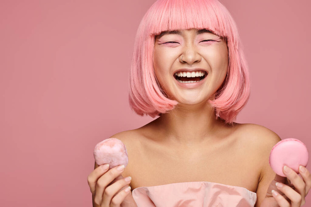 ελκυστική Ασιάτισσα στα 20 με ροζ μαλλιά χαρούμενη γελώντας και κρατώντας γλυκά σε ζωντανό φόντο - Φωτογραφία, εικόνα