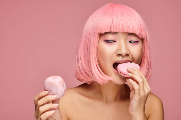 linda asiático jovem mulher com rosa cabelo comer doces contra vibrante fundo - Foto, Imagem