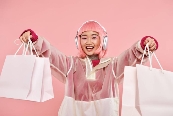 fröhliche Asiatin mit pinkfarbenen Haaren und Einkaufstaschen in Kopfhörern vor lebendigem Hintergrund - Foto, Bild