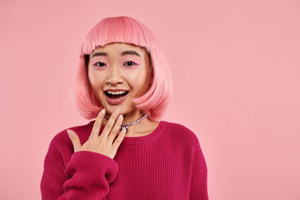 happy asian girl in vibrant sweater with pearl necklace expressing admiration on pink background - Foto, afbeelding