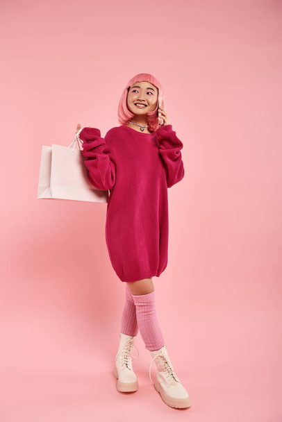 asiático mujer con rosa pelo vocación de smartphone y celebración paquete sobre hombro en rosa fondo - Foto, imagen