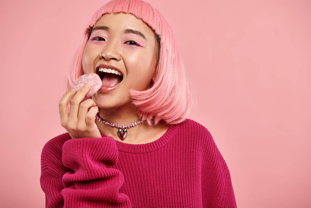 retrato de alegre asiático mulher com rosa cabelo comendo mochi com admiração no vibrante fundo - Foto, Imagem