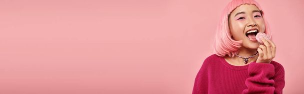 visão horizontal de mulher asiática com cabelo rosa comendo mochi com admiração no fundo vibrante - Foto, Imagem