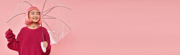 horizontal shot of cute asian woman standing under umbrella and looking to side on pink background - Photo, Image