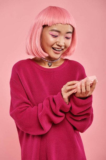cheerful asian young woman looking of gift with admiration against pink background - Photo, Image