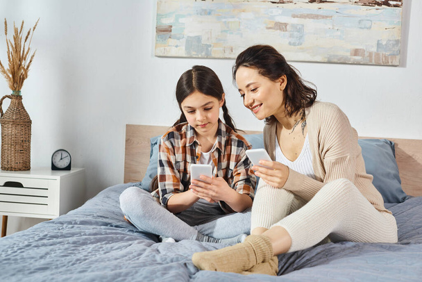 Mutter und Tochter sitzen auf einem Bett, in ein Mobiltelefon vertieft und teilen einen Moment der Verbindung. - Foto, Bild