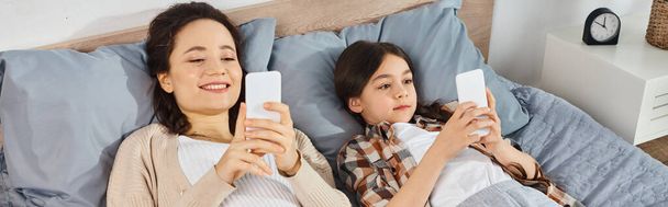 Una mujer y una chica yacen en una cama, enfocadas en sus teléfonos celulares, disfrutando de un tiempo de calidad juntos en casa. - Foto, Imagen