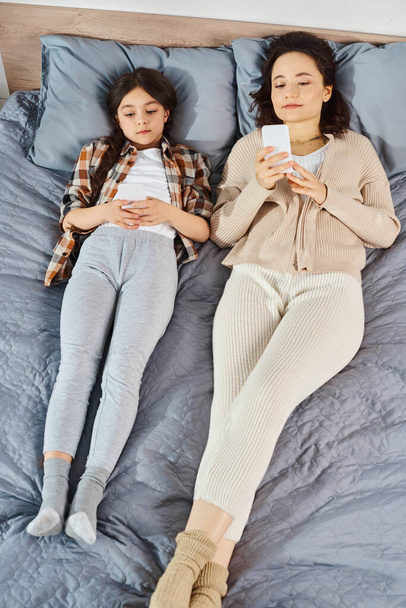 Μια μητέρα και η κόρη είναι ξαπλωμένες σε ένα κρεβάτι, απορροφούνται στα smartphones τους, απολαμβάνοντας ποιοτικό χρόνο μαζί στο σπίτι. - Φωτογραφία, εικόνα